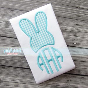 Puede incluir: Tela blanca con un diseño de orejas de conejo a cuadros azul claro y un monograma. Las orejas de conejo y el monograma están delineados en azul claro. El texto "pick & stitch applique designs" está en la parte inferior.