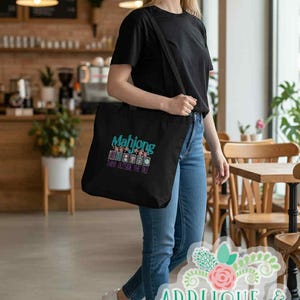 K&ouml;nnte beinhalten: Eine schwarze Tragetasche mit dem Wort "Mahjong" in T&uuml;rkis und Pink sowie dem Satz "Think Outside The Tile". Die Tasche wird &uuml;ber der Schulter getragen. Die Person tr&auml;gt ein schwarzes T-Shirt und blaue Jeans.