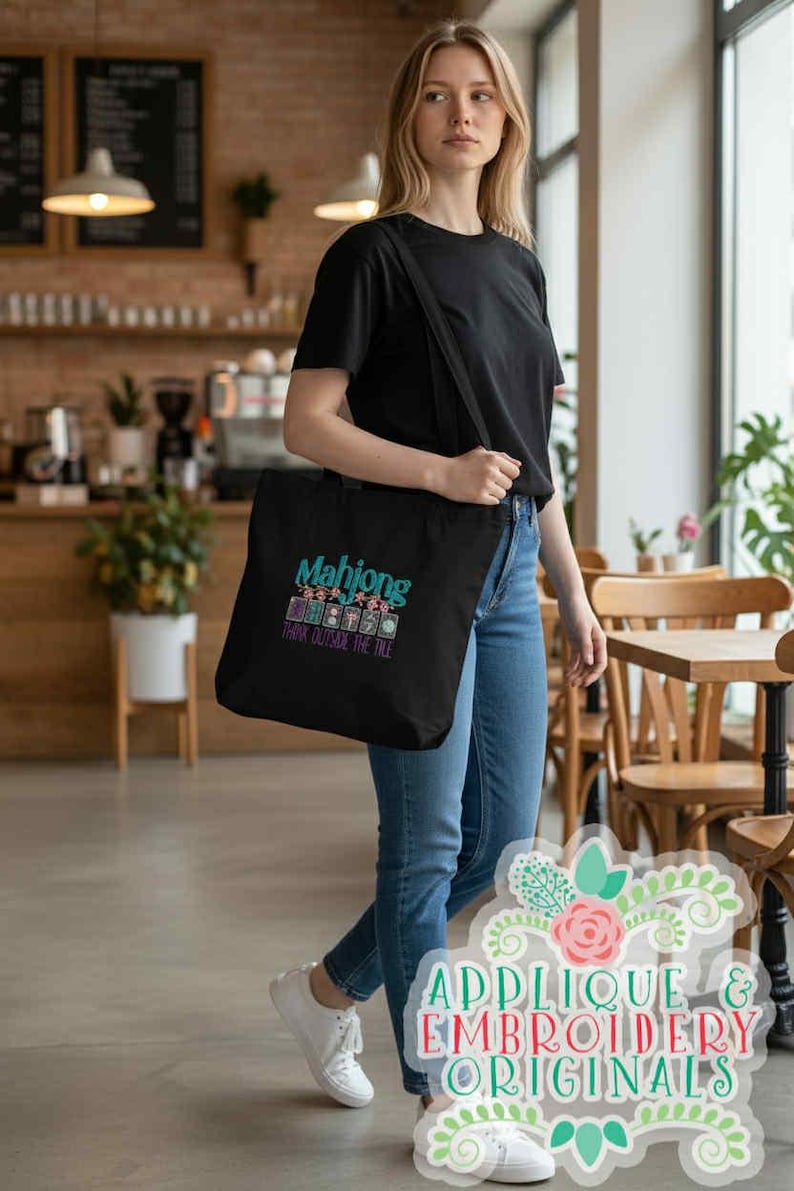 K&ouml;nnte beinhalten: Eine schwarze Tragetasche mit dem Wort "Mahjong" in T&uuml;rkis und Pink sowie dem Satz "Think Outside The Tile". Die Tasche wird &uuml;ber der Schulter getragen. Die Person tr&auml;gt ein schwarzes T-Shirt und blaue Jeans.