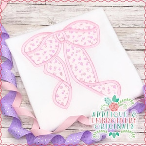 May include: A pink and white floral applique design of a bow. The applique is on a white fabric background. The text "Applique & Embroidery Originals" is in the lower right corner of the image.