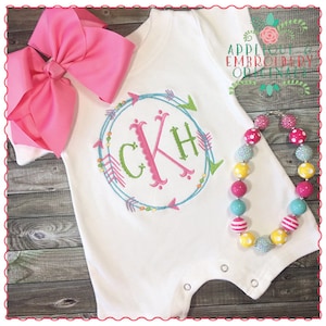 May include: White baby onesie with a pink, green, and blue embroidered monogram in a circle with arrows. The monogram is "CKH".