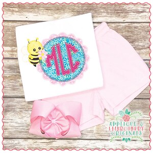May include: A white shirt with a pink and blue floral circle applique featuring a bee and the monogram "MLL". The shirt is paired with pink shorts and a pink bow.