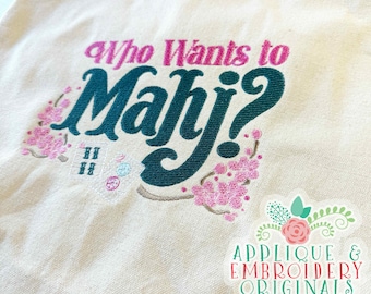 May include: A beige tote bag with the embroidered text "Who Wants to Mahj?" in teal and pink. The design includes pink floral accents and a small sticker with text and floral graphics.