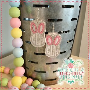 Puede incluir: Un par de pendientes blancos en forma de conejo con orejas rosas y un monograma en el centro. Los pendientes cuelgan de un cubo de metal con una guirnalda de cuentas de colores pastel en primer plano. El texto "Applique & Embroidery Originals" es visible en la esquina inferior derecha de la imagen.