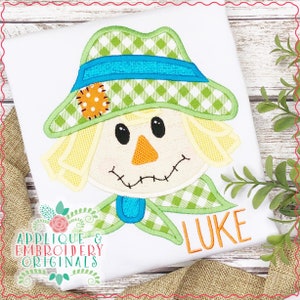 May include: A green and yellow appliqué of a scarecrow's face with a blue and green checkered hat. The name "LUKE" is embroidered in orange below the scarecrow.