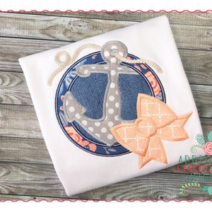 May include: A white t-shirt with an embroidered design of a grey anchor with white polka dots, a blue and white circle, and a peach bow. The design is surrounded by a rope.
