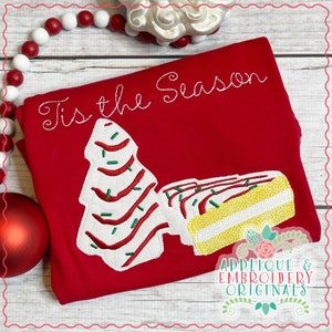 May include: Red t-shirt with white embroidery of a Christmas tree and a cake with the text "Tis the Season".