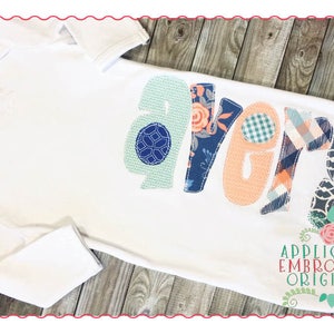 May include: A white baby gown with the name "Avery" spelled out in colorful fabric letters. The letters are appliqued with different patterns, including floral, plaid, and geometric designs. The gown is on a wooden background with the text "Applique & Embroidery Originals" at the bottom.