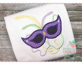 May include: Purple and black Mardi Gras mask with gold and green beads and feathers. The mask is embroidered on a white fabric.