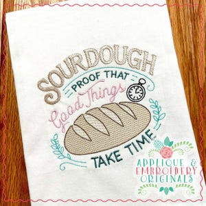 Puede incluir: Una toalla de cocina blanca con un diseño bordado que dice "Sourdough - Proof That Good Things Take Time". El diseño presenta una hogaza de pan, un reloj y ramitas con hojas.