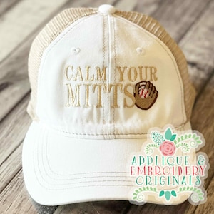May include: White baseball cap with a tan mesh back. The cap features the embroidered phrase "CALM YOUR MITTS" in tan thread, along with a baseball glove and ball design. A sticker with the text "APPLIQUE & EMBROIDERY ORIGINALS" is on the right side.