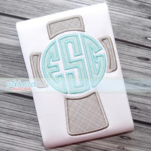 Puede incluir: Un artículo de tela blanca con un diseño de monograma en forma de cruz. El monograma es de color azul claro, y los brazos de la cruz son de color marrón claro con un patrón texturizado. El texto "pick & stitch applique designs" está en la esquina inferior izquierda.