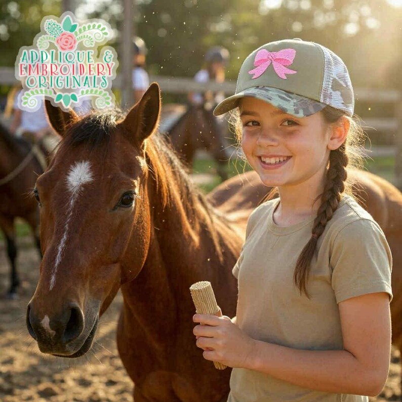May include: A young person wearing a camouflage baseball cap with a pink bow, smiling next to a brown horse. The cap has a white mesh back. The image includes the text "Applique & Embroidery Originals".