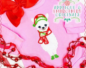 May include: A pink baby onesie with a white llama wearing a red and green plaid scarf and a green hat. The llama is embroidered on the onesie. The onesie is surrounded by red beads and a red ribbon.