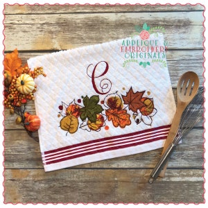 May include: White kitchen towel with a red and white striped border. The towel features an embroidered design of fall leaves in shades of orange, yellow, green, and brown. The letter "C" is embroidered in the upper left corner. The text "Applique & Embroidery Originals" is embroidered in the upper right corner.