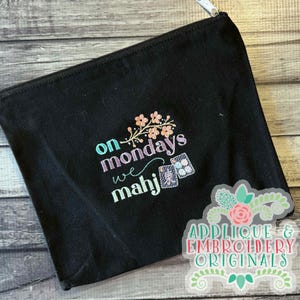 May include: Black zippered pouch with embroidered text "on mondays we mahj" in teal, purple, and green, with a floral accent and mahjong tile graphics. The pouch has a silver zipper and a decorative sticker in the lower right corner.