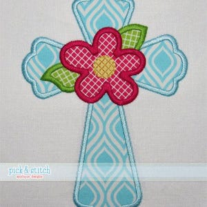 Puede incluir: Aplicación de cruz bordada con una flor rosa y hojas verdes. La cruz es azul claro con un patrón blanco y azul, delineado en azul más oscuro. La flor tiene un centro amarillo y un patrón de cuadrícula blanca.