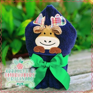 Op de afbeelding: Een blauwe badhanddoek met capuchon met een bruine eland-applicatie op de capuchon. De handdoek is vastgebonden met een groene strik. De tekst "Applique & Embroidery Originals" is zichtbaar in de linkerbenedenhoek van de afbeelding.
