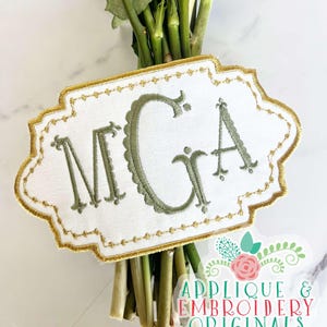May include: A white fabric flower bouquet tag with a gold border and green embroidered letters spelling "MGA".
