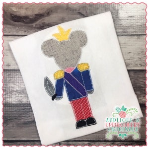 May include: A gray mouse dressed as a toy soldier with a yellow crown, blue jacket, purple sash, red pants, and a silver sword. The mouse is embroidered on a white t-shirt.