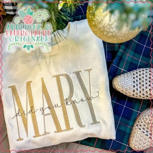 Puede incluir: Una sudadera blanca con el texto "MARY did you know?" bordado en hilo dorado y plateado.