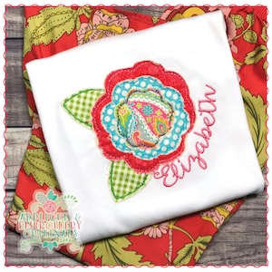 May include: White t-shirt with a large, colorful floral applique. The applique features a paisley design in the center, surrounded by layers of red, blue, and green. The name "Elizabeth" is embroidered in pink script. The shirt rests on a red floral patterned fabric.