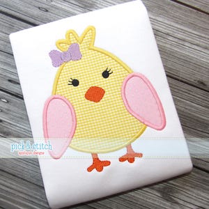 Puede incluir: Tela blanca con un diseño de pollito amarillo bordado. El pollito tiene un cuerpo a cuadros amarillo y blanco, alas rosas y un lazo morado. El pollito tiene patas y pico naranjas y ojos negros. El texto "pick & stitch applique designs" está en la parte inferior.