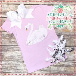 May include: A pink t-shirt with a white swan applique. The swan is embroidered with a sparkly design. The shirt is on a wooden background with white and silver ribbon.