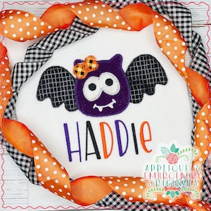 Puede incluir: Un pelele blanco con una aplicación de murciélago morado con alas negras y un lazo amarillo. El nombre "Haddie" está bordado en letras moradas y negras debajo del murciélago. El pelele está rodeado de una cinta a cuadros blanco y negro con lunares naranjas.