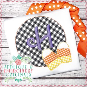 May include: White t-shirt with a black and white checkered circle design. The circle contains a purple embroidered letter "H" and two candy corn appliques. An orange polka dot ribbon is in the background. The text "Applique & Embroidery Originals" is at the bottom.