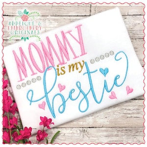 Puede incluir: Camiseta blanca bordada con el texto "Mommy is my bestie" en letras rosas, azules y doradas. La camiseta también presenta pequeños corazones rosas y azules.
