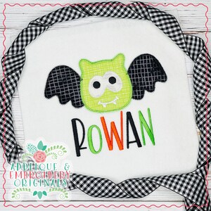 Puede incluir: Babero blanco para bebé con un aplique de murciélago verde y el nombre "ROWAN" en negro, verde y naranja. Una cinta a cuadros en blanco y negro enmarca el babero. También se ven las palabras "Applique & Embroidery Originals".