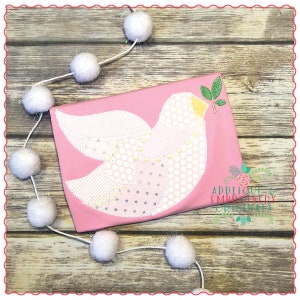 May include: A pink t-shirt featuring a white appliqué of a dove with a green leaf in its beak. The dove is crafted from various patterned fabrics, including polka dots, stripes, and floral prints. The t-shirt is displayed against a wooden backdrop with a white pom-pom garland.