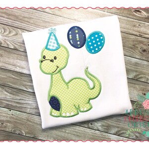May include: Green dinosaur wearing a blue and white party hat and holding two balloons, one blue with white polka dots and one blue with green and white polka dots. The dinosaur is on a white shirt.