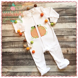 May include: White baby onesie with a zipper down the front. The onesie has an embroidered pumpkin design in orange, green, and brown.