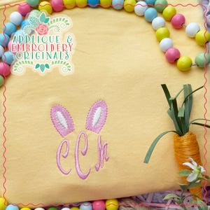 Peut inclure: Un t-shirt jaune avec des oreilles de lapin roses et le monogramme "CCH" brodé sur le devant. Le t-shirt est entouré d'une bordure de perles colorées.