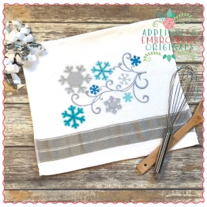 Puede incluir: Una toalla de cocina blanca con un borde gris y un diseño bordado de copos de nieve y una vid. El diseño está en tonos de azul y gris.