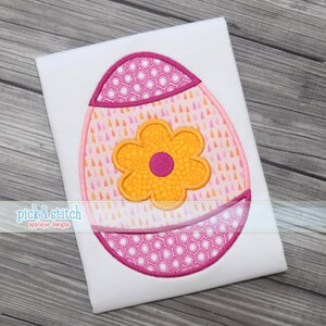 Puede incluir: Un diseño de aplique de huevo de Pascua sobre tela blanca. El huevo está decorado con patrones rosas y naranjas, una flor amarilla y un borde rosa. El texto "pick & stitch applique designs" está en la esquina inferior izquierda.