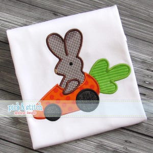 Puede incluir: Tela blanca con un diseño de aplique que presenta un conejito en un coche zanahoria. El conejito tiene un patrón a cuadros marrón y blanco. El coche zanahoria es naranja con ruedas negras y hojas verdes. El texto "pick & stitch applique designs" está en la parte inferior.