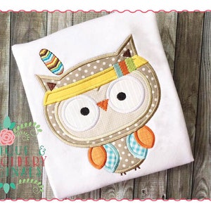 May include: Embroidered design of a brown owl with yellow, orange, blue, and white accents. The owl is wearing a yellow headband with a feather on top. The design is on a white t-shirt.