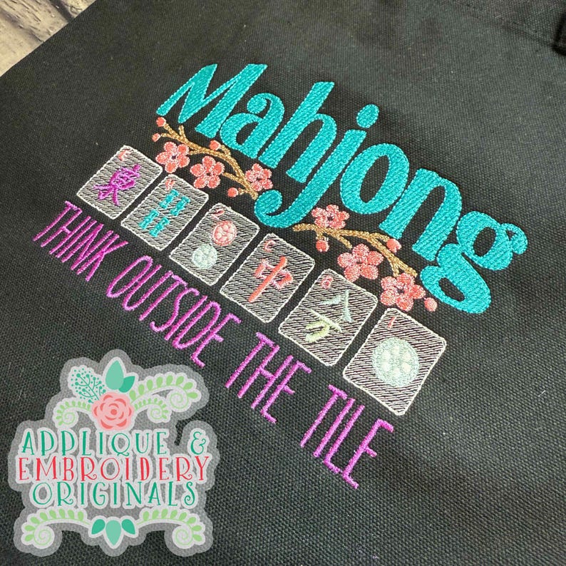 K&ouml;nnte beinhalten: Schwarze Tragetasche mit gesticktem Text und Grafiken. Das Wort "Mahjong" ist in T&uuml;rkis gehalten, mit rosa floralen Akzenten. Darunter steht der Satz "THINK OUTSIDE THE TILE" in Lila. Die Tasche hat auch gestickte Mahjong-Kacheln.