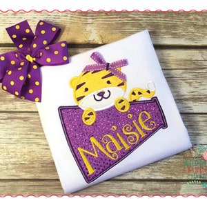 May include: A white t-shirt with a purple and yellow embroidered design of a tiger holding a purple and yellow megaphone with the name "Maisie" on it. A purple and yellow polka dot bow is on the top left of the shirt.