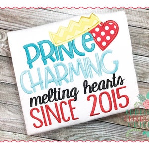 May include: White t-shirt with embroidered text that reads "Prince Charming melting hearts since 2015". The text is in blue, red, and black. There is a yellow crown and a red heart with white polka dots above the text.