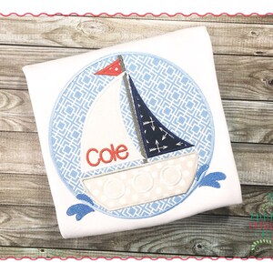 May include: A white t-shirt with a blue and white sailboat design in a circle. The sailboat has a red flag and the name "Cole" is embroidered on the sail.