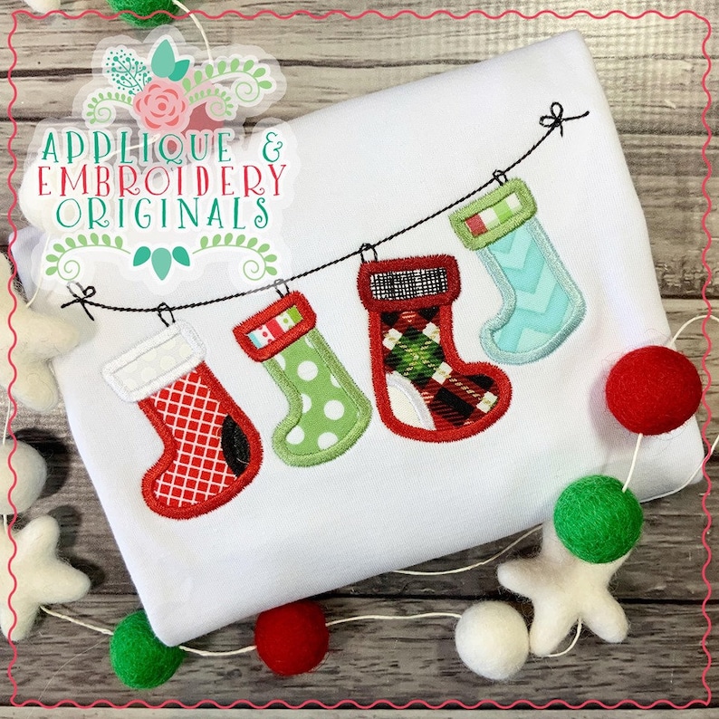 May include: A white shirt with a string of embroidered Christmas stockings hanging from it. The stockings are red, green, and blue with different patterns. The shirt is surrounded by white, red, and green pom-poms.