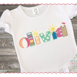May include: White t-shirt with a colorful embroidered name "Olivia" with a beach theme. The letters are decorated with stripes, polka dots, and a sun.