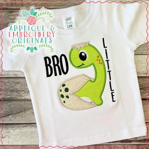 May include: White t-shirt with a green dinosaur applique and the text "BRO" and "LITTLE" in black embroidery.