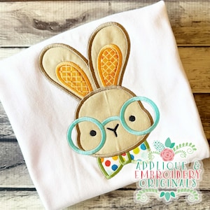 Puede incluir: Una camiseta blanca con una aplicación bordada de una cabeza de conejo color canela con orejas largas, interiores con patrón naranja y gafas azul claro. El conejo lleva una pajarita blanca con lunares de colores. El texto dice Applique and Embroidery Originals.