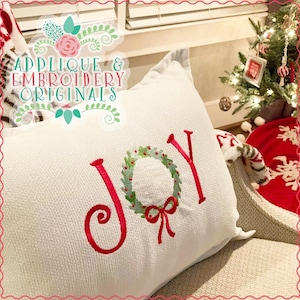 May include: A white textured pillow with the embroidered word "JOY" in red. The "O" is a wreath with green leaves and red berries. The pillow is part of a Christmas theme.