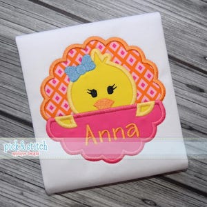 Puede incluir: Un artículo de tela blanca con un diseño bordado de un pollito amarillo con un lazo azul, dentro de un marco festoneado rosa y naranja. El nombre "Anna" está bordado en amarillo sobre una pancarta rosa en forma de nube.